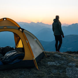 Woman standing on a cliff overlooking a sunset next to her tent.